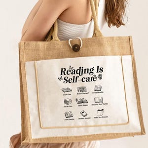 May include: A beige jute tote bag with a white pocket featuring the text "Reading Is Self-care" and illustrations of books. The bag has a button closure and tan handles.