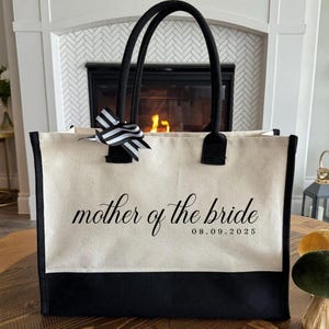 Canvas tote bag featuring the text “Mother of the Bride” in elegant script with a custom date printed underneath; designed on a two-tone cotton canvas tote with sturdy handles and a ribbon accent; displayed in a neutral soft-lit background; perfect personalized wedding gift for mothers, brides, or bridal parties.