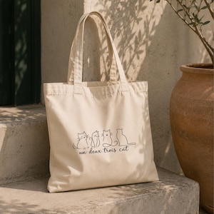 May include: A cream-coloured canvas tote bag featuring a black line-art graphic of four cats sitting in a row. Below the cats, the text reads "un deux trois cat" in a simple, dark script font. The bag has two matching fabric shoulder straps.