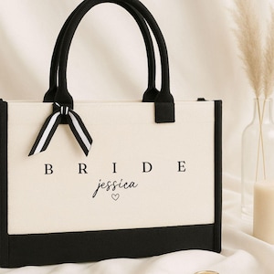 May include: A rectangular canvas tote bag in cream with black trim and black handles. The front features the word BRIDE in a serif font above the name jessica in a script font with a small heart. A black and white striped ribbon is tied to one handle.