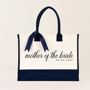 Canvas tote bag featuring the text “Mother of the Bride” in elegant script with a custom date printed underneath; designed on a two-tone cotton canvas tote with sturdy handles and a ribbon accent; displayed in a neutral soft-lit background; perfect personalized wedding gift for mothers, brides, or bridal parties.