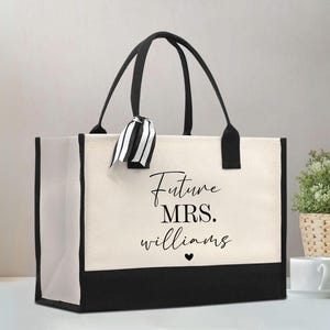 May include: A rectangular canvas tote bag with a cream-colored body and black base, side panels, and handles. The front features the black text "Future MRS. williams" with a small heart icon below. A black and white striped ribbon is tied to one handle.