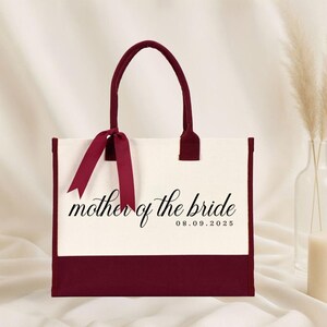 Canvas tote bag featuring the text “Mother of the Bride” in elegant script with a custom date printed underneath; designed on a two-tone cotton canvas tote with sturdy handles and a ribbon accent; displayed in a neutral soft-lit background; perfect personalized wedding gift for mothers, brides, or bridal parties.