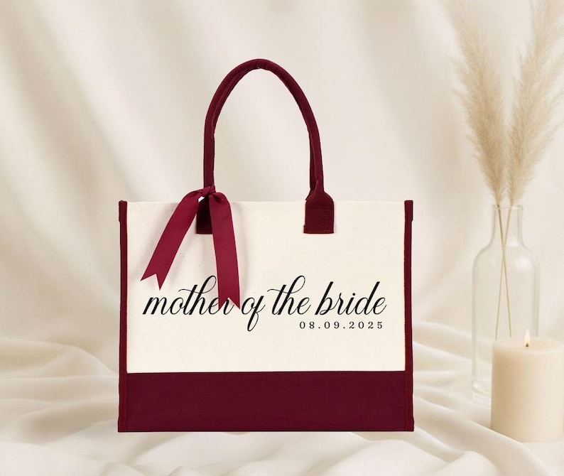 Canvas tote bag featuring the text “Mother of the Bride” in elegant script with a custom date printed underneath; designed on a two-tone cotton canvas tote with sturdy handles and a ribbon accent; displayed in a neutral soft-lit background; perfect personalized wedding gift for mothers, brides, or bridal parties.