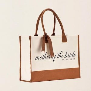 Canvas tote bag featuring the text “Mother of the Bride” in elegant script with a custom date printed underneath; designed on a two-tone cotton canvas tote with sturdy handles and a ribbon accent; displayed in a neutral soft-lit background; perfect personalized wedding gift for mothers, brides, or bridal parties.