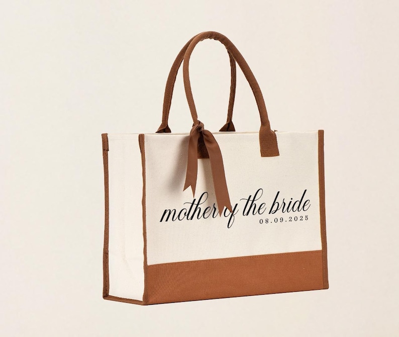 Canvas tote bag featuring the text “Mother of the Bride” in elegant script with a custom date printed underneath; designed on a two-tone cotton canvas tote with sturdy handles and a ribbon accent; displayed in a neutral soft-lit background; perfect personalized wedding gift for mothers, brides, or bridal parties.