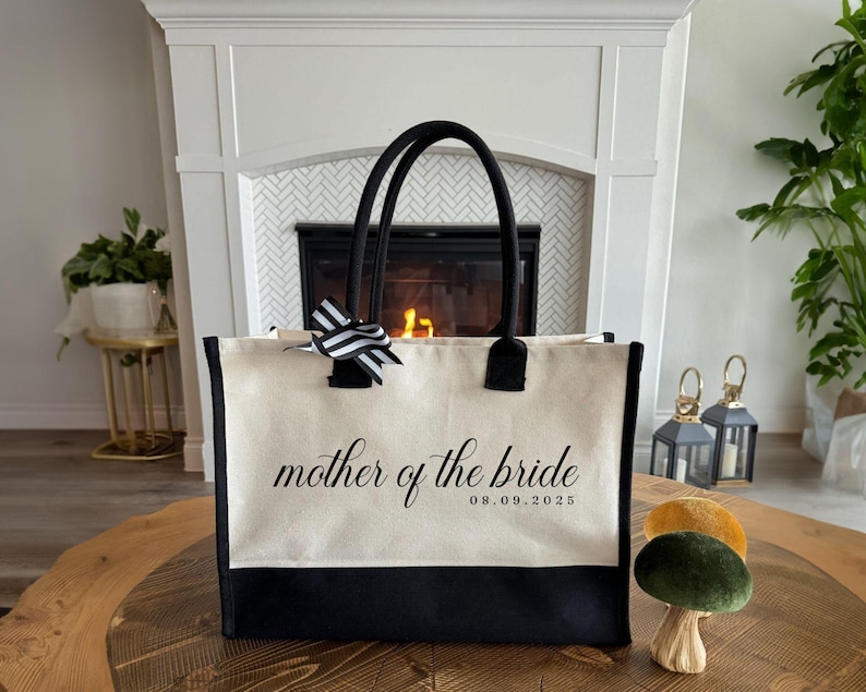 Canvas tote bag featuring the text “Mother of the Bride” in elegant script with a custom date printed underneath; designed on a two-tone cotton canvas tote with sturdy handles and a ribbon accent; displayed in a neutral soft-lit background; perfect personalized wedding gift for mothers, brides, or bridal parties.