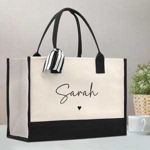 May include: A rectangular canvas tote bag with a cream-colored body and a solid black base. The bag features black handles and a black and white striped ribbon tied to one handle. The name Sarah is printed in black script with a small heart below.