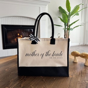 Canvas tote bag featuring the text “Mother of the Bride” in elegant script with a custom date printed underneath; designed on a two-tone cotton canvas tote with sturdy handles and a ribbon accent; displayed in a neutral soft-lit background; perfect personalized wedding gift for mothers, brides, or bridal parties.