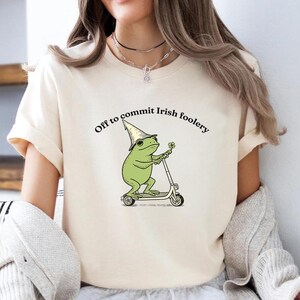 May include: Cream-colored t-shirt featuring a green frog wearing a wizard hat and riding a scooter. The text "Off to commit Irish foolery" is arched above the frog illustration. The design is printed on a short-sleeved tee.