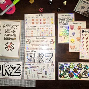 Op de afbeelding: Een verzameling spaarbinders en kaarten met verschillende ontwerpen en tekst, waaronder "STRAY KIDS SAVINGS BINDER" en "SKZ". De kaarten bevatten illustraties, tekst en spaardoelen, met een vijf dollar biljet op de achtergrond.