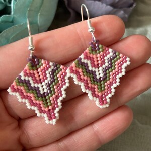 May include: Handmade beaded earrings in a geometric diamond shape. The earrings feature a pattern of pink, white, green, and purple beads. The earrings have silver-colored ear wires and are displayed against a soft background.