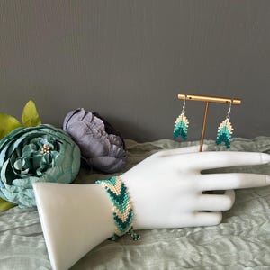 May include: A beaded bracelet and matching earrings set. The bracelet and earrings feature a chevron pattern in shades of teal, white, and green. The jewelry is displayed on a white mannequin hand and a gold-colored earring stand.