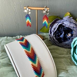May include: A beaded bracelet and earrings set. The bracelet and earrings feature a chevron pattern in shades of red, orange, yellow, green, and blue. The earrings have silver flower studs. The bracelet is displayed on a white stand.