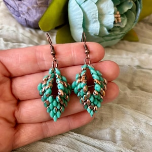 May include: A pair of handmade earrings featuring a leaf-like design. The earrings are composed of small, turquoise-colored beads and bronze-colored beads, with bronze-colored ear wires. The earrings are held in a hand against a soft, blurred background.