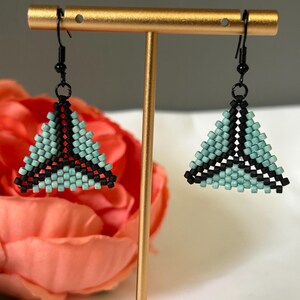 May include: A pair of beaded triangle earrings. The earrings feature a turquoise and black color scheme. The triangles are suspended from black ear wires and are displayed on a gold earring stand. The earrings are a geometric design.
