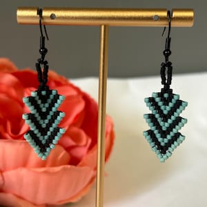 May include: A pair of handmade beaded earrings. The earrings feature a chevron design in black and turquoise. The earrings are suspended from black ear wires and are displayed on a gold earring stand.