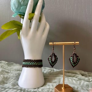 Könnte beinhalten: Ein handgefertigtes Schmuckset mit Armband und Ohrringen. Armband und Ohrringe haben ein gewebtes Design mit braunen und hellgrünen Perlen. Die Ohrringe werden auf einem goldfarbenen Ständer präsentiert, und das Armband auf einer weißen handförmigen Halterung.