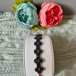 May include: A beaded bracelet with dark blue and copper-colored beads is displayed on a white stand. The bracelet has a floral design. Two large, artificial flowers, one teal and one pink, are in the background.