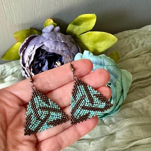 May include: Triangular beaded earrings in turquoise and brown. The earrings have a geometric pattern and are suspended from a bronze-colored hook. The earrings are displayed against a backdrop of flowers and fabric.