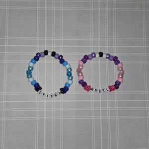 May include: Two beaded bracelets. One is blue and purple with the word "STITCH" spelled out in white letter beads. The other is pink and purple with the word "ANGEL" spelled out in white letter beads. The bracelets are on a gray and white checkered surface.