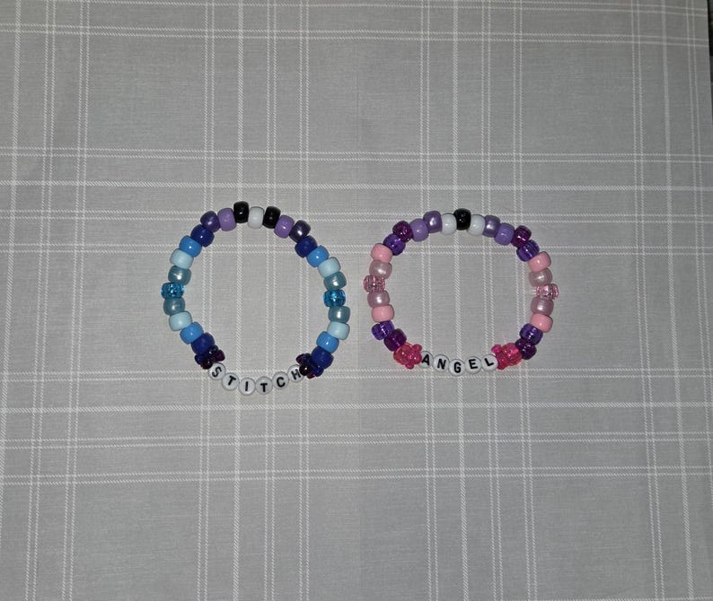 May include: Two beaded bracelets. One is blue and purple with the word "STITCH" spelled out in white letter beads. The other is pink and purple with the word "ANGEL" spelled out in white letter beads. The bracelets are on a gray and white checkered surface.