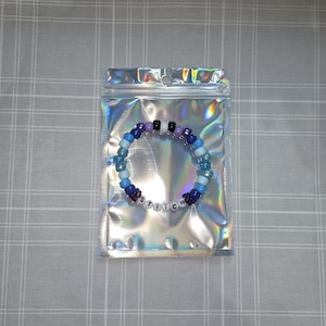 May include: A colorful beaded bracelet in a holographic bag. The bracelet features a mix of blue, purple, black, and white beads, spelling out "STITCH". The bag has a zipper closure and a shiny, iridescent finish.