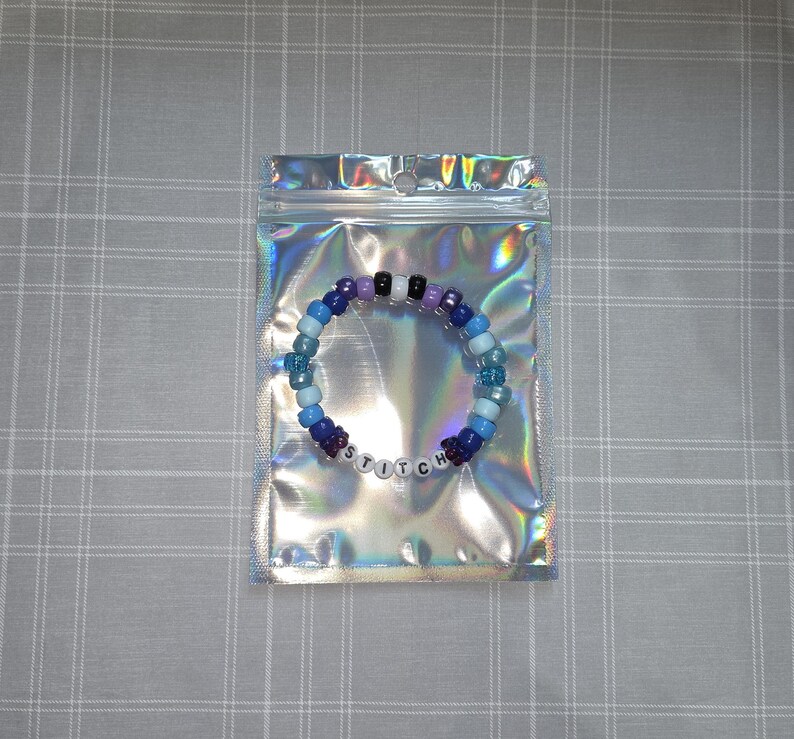 May include: A colorful beaded bracelet in a holographic bag. The bracelet features a mix of blue, purple, black, and white beads, spelling out "STITCH". The bag has a zipper closure and a shiny, iridescent finish.