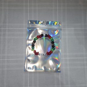 May include: A beaded bracelet spelling "LILO" in white letter beads. The bracelet has red, green, black, and white beads. It is presented inside a silver holographic zip-lock bag.