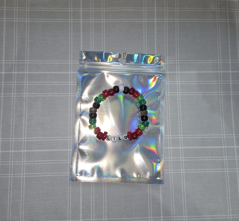 May include: A beaded bracelet spelling "LILO" in white letter beads. The bracelet has red, green, black, and white beads. It is presented inside a silver holographic zip-lock bag.