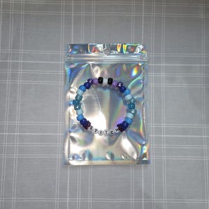May include: A beaded bracelet in a holographic bag. The bracelet has blue, purple, black, and white beads. The word "STITCH" is spelled out with white letter beads. The bag has a zip closure and is iridescent.