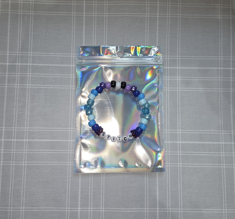 May include: A beaded bracelet in a holographic bag. The bracelet has blue, purple, black, and white beads. The word "STITCH" is spelled out with white letter beads. The bag has a zip closure and is iridescent.
