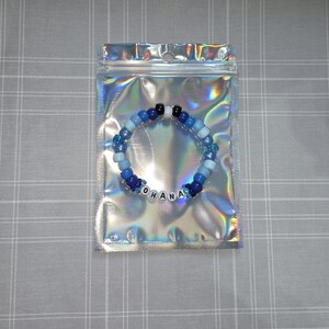 May include: A blue and white beaded bracelet with the word "OHANA" spelled out in white letter beads. The bracelet is in a clear, iridescent plastic bag on a grey and white checkered surface.