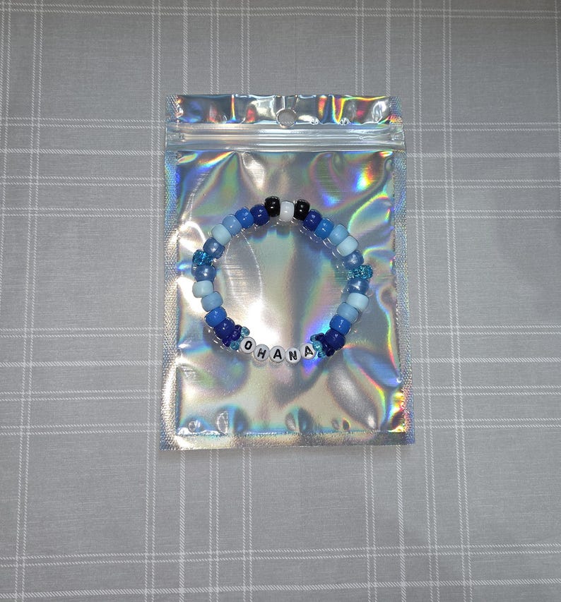 May include: A blue and white beaded bracelet with the word "OHANA" spelled out in white letter beads. The bracelet is in a clear, iridescent plastic bag on a grey and white checkered surface.