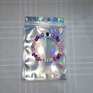 May include: A beaded bracelet spelling "ANGEL" in white letter beads, with pink, purple, and black beads. The bracelet is inside a holographic, iridescent zip-top bag, presented on a grey checkered surface.