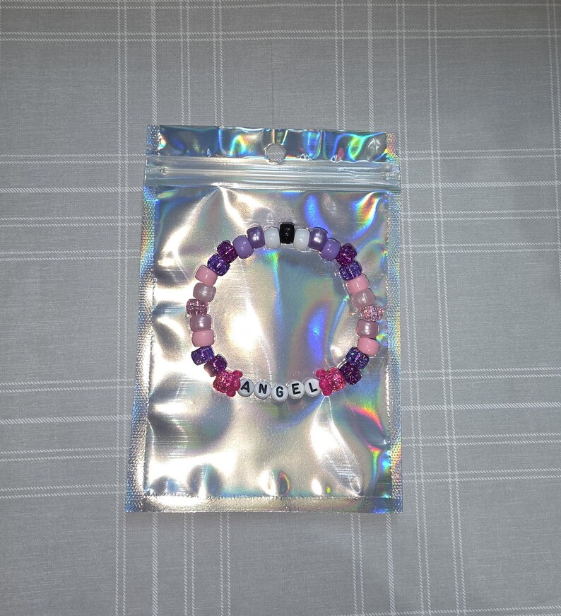 May include: A beaded bracelet spelling "ANGEL" in white letter beads, with pink, purple, and black beads. The bracelet is inside a holographic, iridescent zip-top bag, presented on a grey checkered surface.