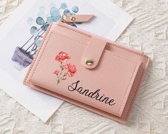 Può includere: Un portafoglio rosa con un motivo floreale e il nome "Sandrine" stampato sul davanti. Il portafoglio ha una chiusura a scatto e una cerniera. Lo sfondo include un acquerello di una pianta.