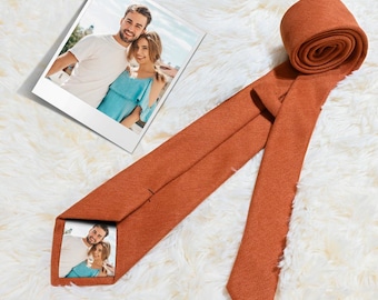 Puede incluir: Una corbata naranja con un extremo enrollado, con una impresión fotográfica en el interior. Una foto enmarcada de una pareja se coloca junto a la corbata. La corbata está hecha de una tela texturizada.