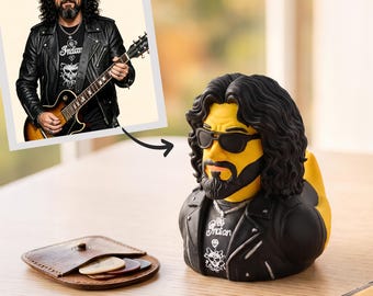 May include: A yellow rubber duck styled as a long-haired man with a beard, wearing black sunglasses, a black leather jacket, and a black t-shirt with the word Indian and a skull graphic. Beside it sits a small brown leather guitar pick holder.