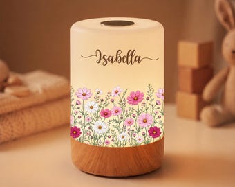 May include: A cylindrical night light with a frosted white top and a wooden base. The light features a floral pattern of pink, magenta, and white cosmos flowers around the bottom, with the name Isabella printed in black script in the centre.
