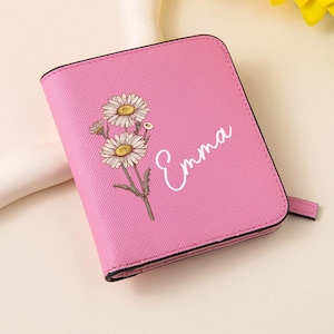 Puede incluir: Cartera rosa con un diseño floral de margaritas y el nombre "Emma" en escritura blanca. La cartera tiene una superficie texturizada y un borde negro. Una pequeña pestaña se extiende desde el lateral.