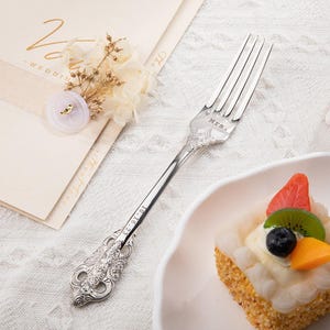 May include: A silver fork with ornate detailing, engraved with "Mrs." and the date "10.16.23". The fork is next to a small cake topped with fruit and a wedding invitation with floral accents.