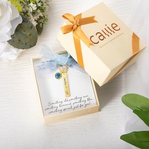 Op de afbeelding: Een goudkleurige sleutelhanger met een blauwe edelsteen en een rechthoekig label met de inscriptie "Jonathan & Sofia" ligt in een crèmekleurige geschenkdoos. De doos is vastgebonden met een gouden lint en bevat de tekst "Something old something new..."