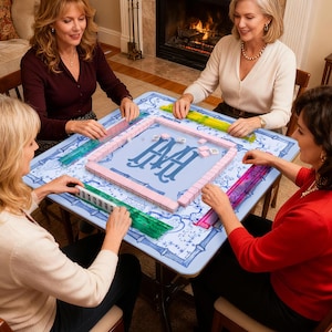 May include: Four women playing Mahjong at a light blue table with a floral design and a monogram. The game pieces are pink and white, and the table has colorful racks. A fireplace is in the background.