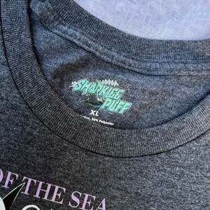 May include: Dark gray XL t-shirt with the text "SHARKIEE PUFF" in a teal and white design. The shirt also features the text "OF THE SEA" in a light purple color. The shirt is made of 50% cotton and 50% polyester.