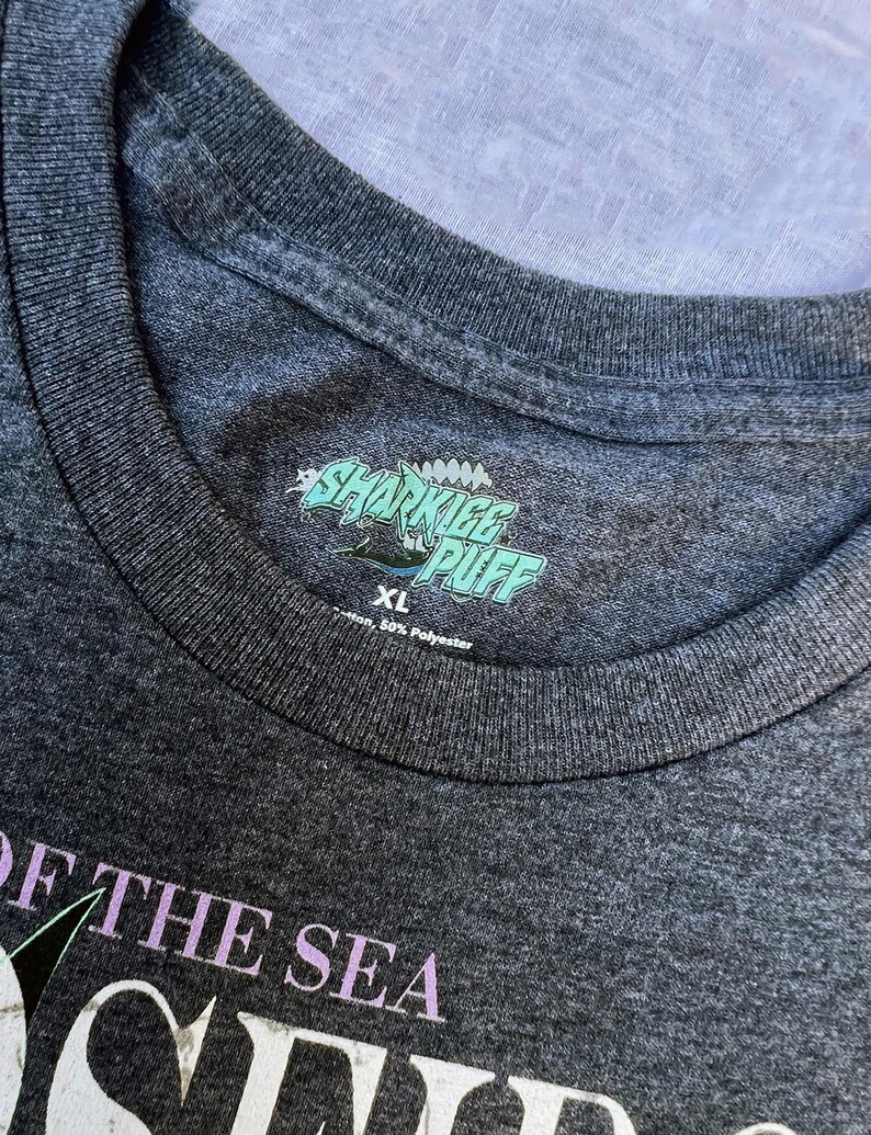 May include: Dark gray XL t-shirt with the text "SHARKIEE PUFF" in a teal and white design. The shirt also features the text "OF THE SEA" in a light purple color. The shirt is made of 50% cotton and 50% polyester.