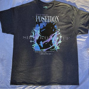 May include: Dark gray t-shirt featuring a graphic of Poseidon, the Greek god of the sea, with the text "POSEIDON" and "GOD OF THE SEA." The design includes blue, purple, and green accents. The shirt also has the text "THESE MORTALS DO CONCERN ME, DIVING AS THEY ARE" and "god of the sea."