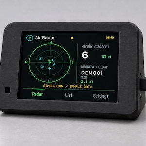 May include: A black rectangular electronic device with a screen displaying an Air Radar interface. The screen shows a circular radar grid with aircraft icons, the text Nearby Aircraft 6, 25 mi, and Nearest Flight DEMO01, 3.1 mi. A power cable is plugged into the side.