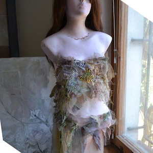 May include: A mannequin bust is adorned with a unique, layered garment. The top features a mix of sheer fabrics, dried leaves, and small yellow floral accents. The skirt portion is a soft, flowing fabric. The bust has long, auburn hair.