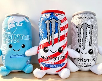 May include: Three plush toys shaped like energy drink cans. The left is light blue with Red Bull Winter Edition branding. The center is red, white, and blue with Monster Energy branding. The right is white and gray with Monster Energy Ultra White branding.